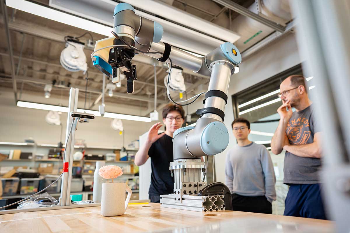 Khoury faculty member Robert Platt, at right, stands next to two other Khoury researchers while watching a robotic arm that is set up on a wooden table reach for a mug