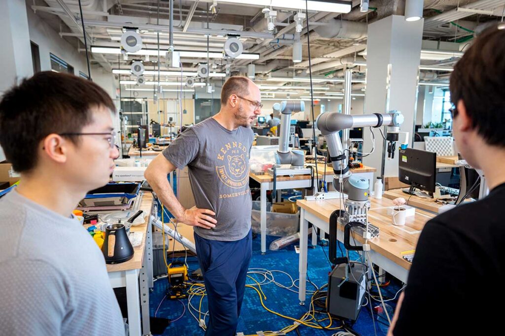 A Khoury researcher stands next to a robotic arm while two students look on